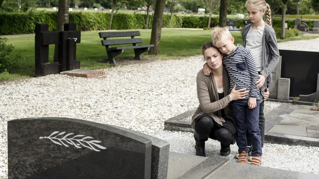 Trauernde Familie am Grab. Die Mutter tröstet ihren Sohn, während die ältere Schwester daneben steht. Sie denken vielleicht an die Zukunft und die halbwaisenrente mindestbetrag.