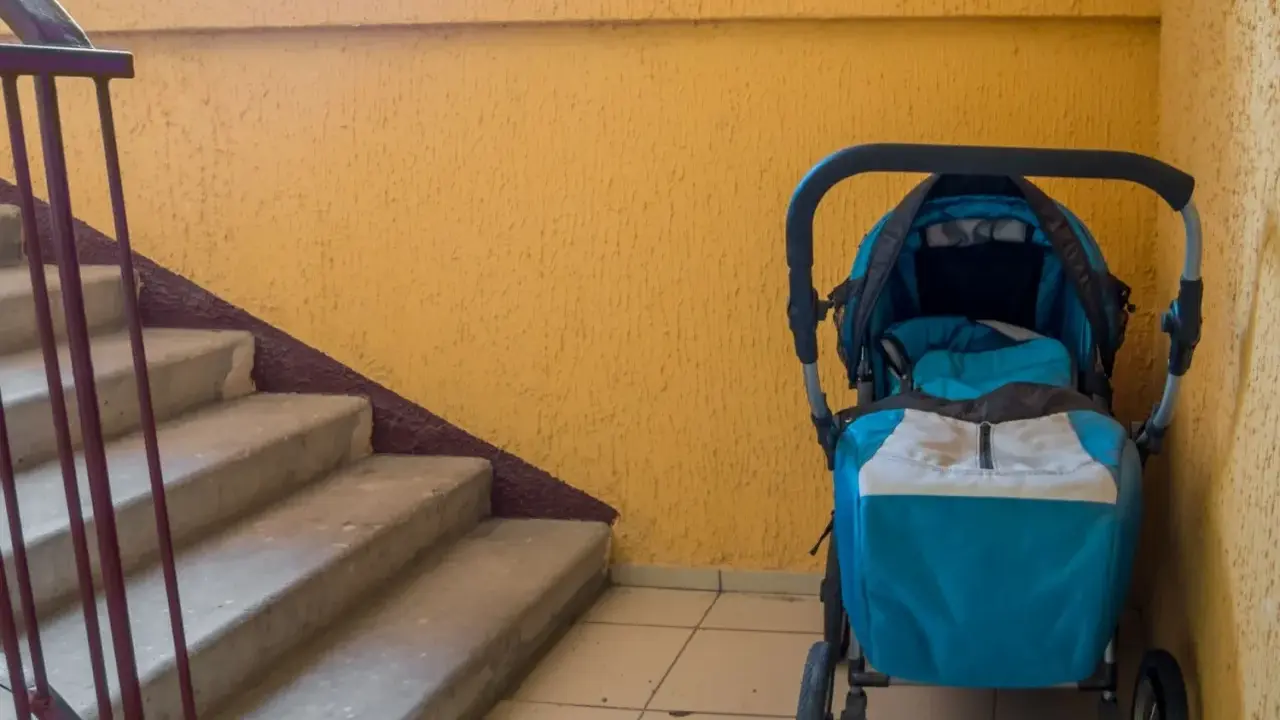 Ein blauer Kinderwagen steht im Treppenhaus neben einer gelben Wand und einer Betontreppe.
