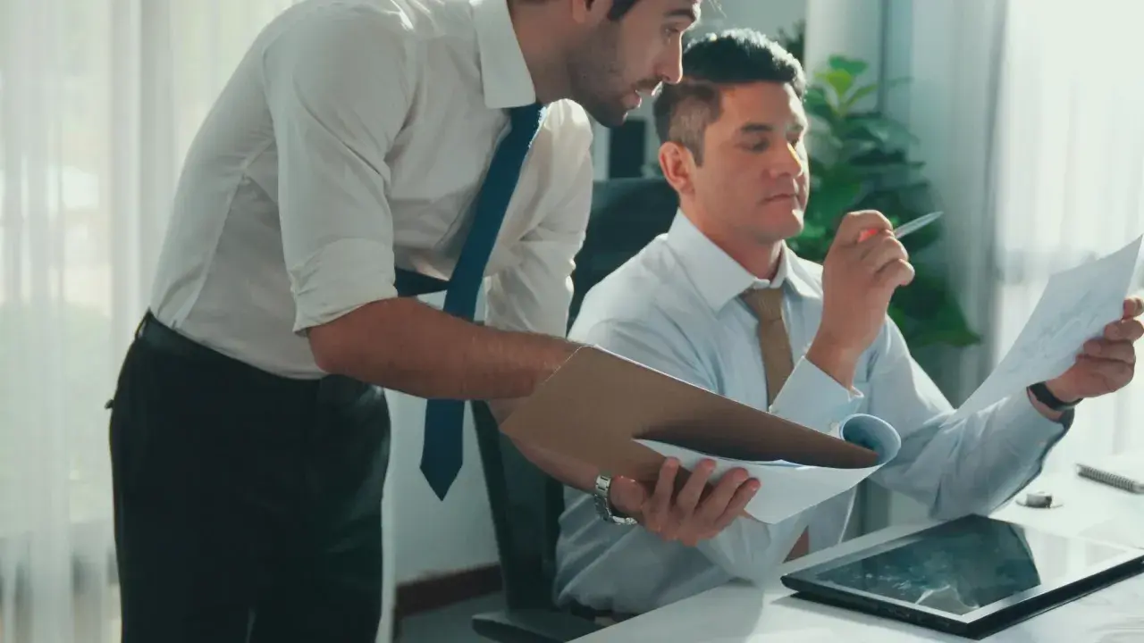 Zwei Männer im Büro besprechen Dokumente. Einer hält eine Mappe, der andere prüft ein Blatt. Vielleicht geht es um bezahlte Freistellung statt Abfindung.