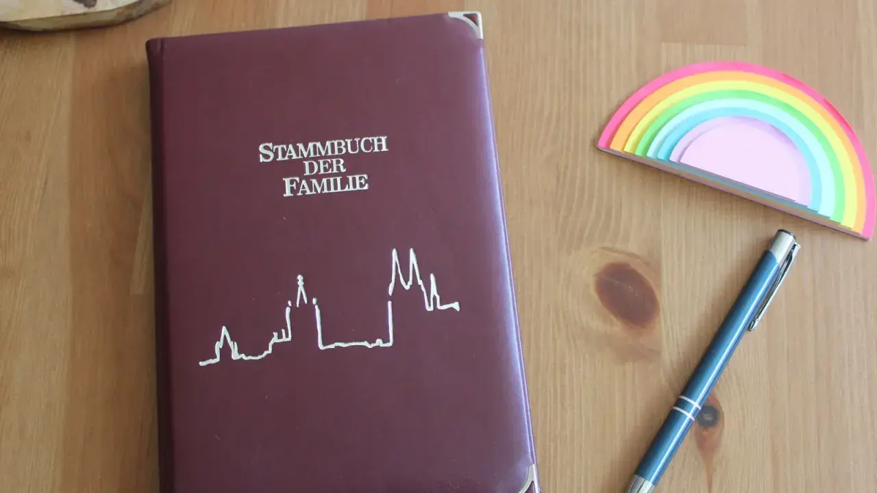 Ein dunkelrotes Stammbuch der Familie liegt neben einem blauen Stift und einem bunten Regenbogen-Notizblock. Hierfür braucht man ein Stammbuch.