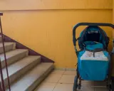 Ein blauer Kinderwagen steht im Treppenhaus neben einer gelben Wand und einer Betontreppe.
