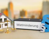 Hausmodell, Waage und Aktenordner mit "Mietminderung" beschriftet. Ein Paragraphenzeichen symbolisiert das Recht bei Mietminderung von Kalt- oder Warmmiete.