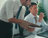 Zwei Männer im Büro besprechen Dokumente. Einer hält eine Mappe, der andere prüft ein Blatt. Vielleicht geht es um bezahlte Freistellung statt Abfindung.