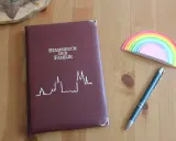 Ein dunkelrotes Stammbuch der Familie liegt neben einem blauen Stift und einem bunten Regenbogen-Notizblock. Hierfür braucht man ein Stammbuch.