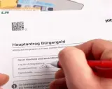 Hand füllt Antrag auf Bürgergeld aus. Geld liegt daneben. Der Antrag betrifft den Selbstbehalt Hartz 4.