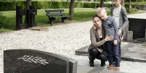 Trauernde Familie am Grab. Die Mutter tröstet ihren Sohn, während die ältere Schwester daneben steht. Sie denken vielleicht an die Zukunft und die halbwaisenrente mindestbetrag.