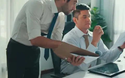 Zwei Männer im Büro besprechen Dokumente. Einer hält eine Mappe, der andere prüft ein Blatt. Vielleicht geht es um bezahlte Freistellung statt Abfindung.