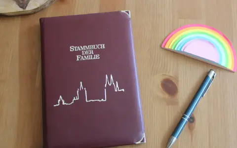 Ein dunkelrotes Stammbuch der Familie liegt neben einem blauen Stift und einem bunten Regenbogen-Notizblock. Hierfür braucht man ein Stammbuch.