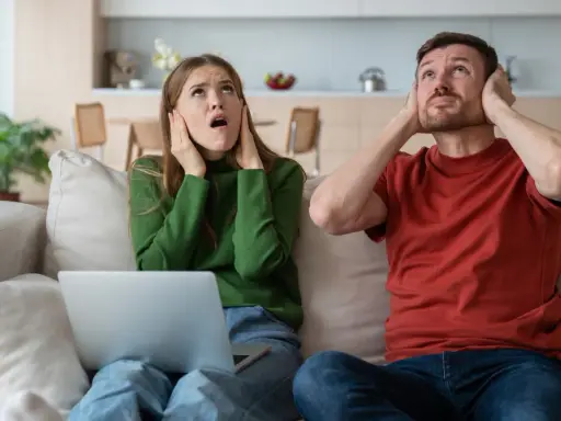 Genervtes Paar auf dem Sofa, Hände an den Ohren. Seit die Mittagsruhe BW abgeschafft ist, ist es hier nie mehr ruhig.