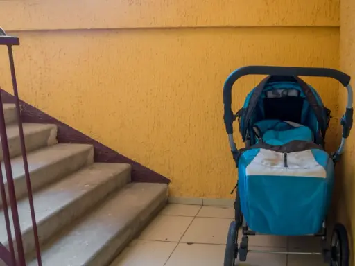Ein blauer Kinderwagen steht im Treppenhaus neben einer gelben Wand und einer Betontreppe.