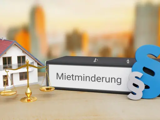 Hausmodell, Waage und Aktenordner mit "Mietminderung" beschriftet. Ein Paragraphenzeichen symbolisiert das Recht bei Mietminderung von Kalt- oder Warmmiete.
