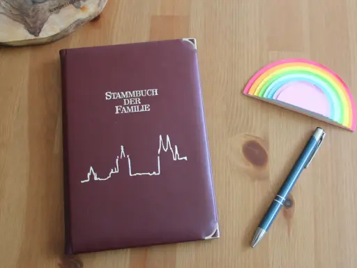 Ein dunkelrotes Stammbuch der Familie liegt neben einem blauen Stift und einem bunten Regenbogen-Notizblock. Hierfür braucht man ein Stammbuch.
