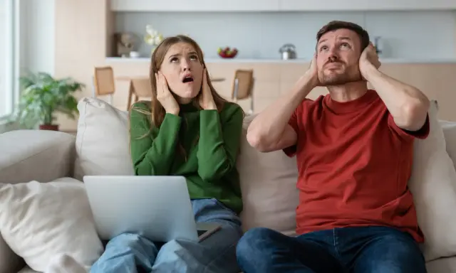 Genervtes Paar auf dem Sofa, Hände an den Ohren. Seit die Mittagsruhe BW abgeschafft ist, ist es hier nie mehr ruhig.