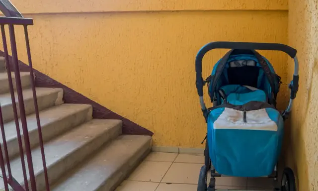 Ein blauer Kinderwagen steht im Treppenhaus neben einer gelben Wand und einer Betontreppe.
