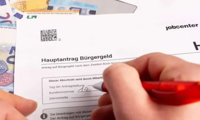 Hand füllt Antrag auf Bürgergeld aus. Geld liegt daneben. Der Antrag betrifft den Selbstbehalt Hartz 4.