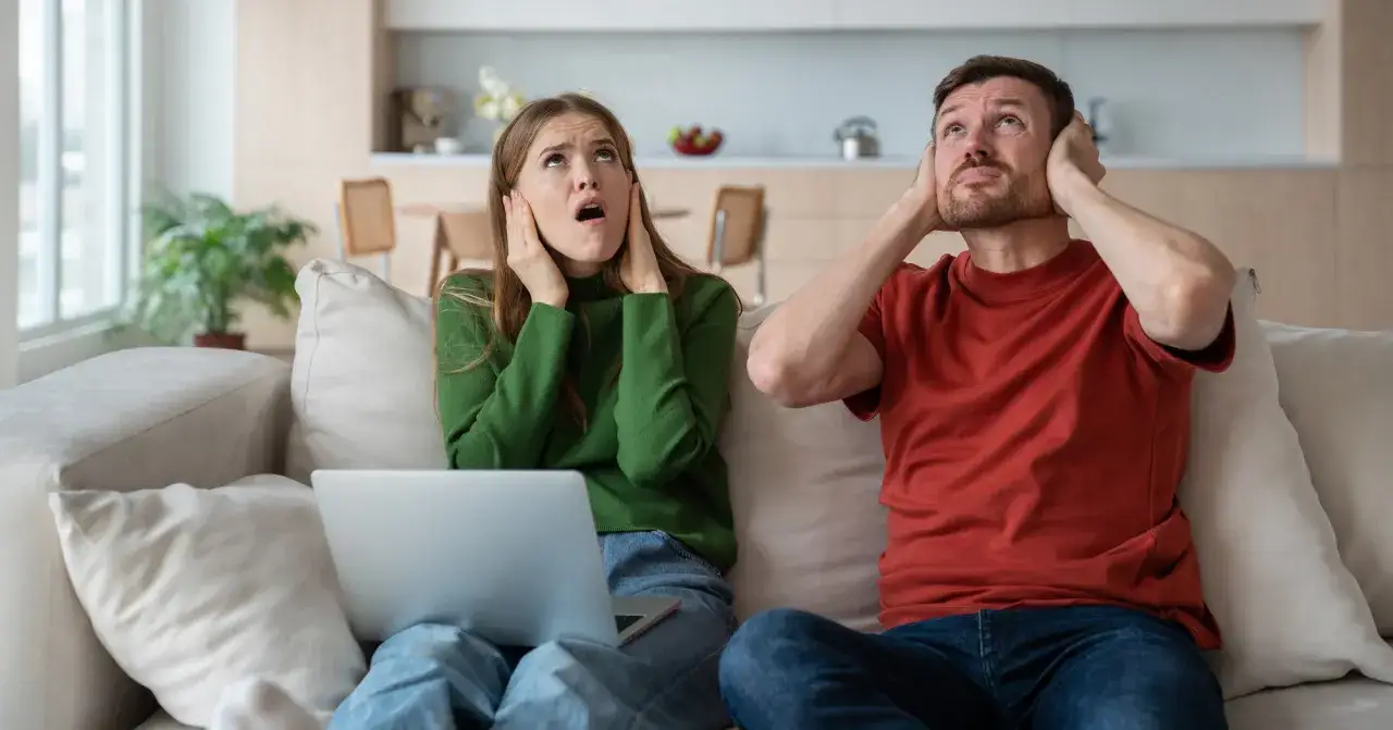 Genervtes Paar auf dem Sofa, Hände an den Ohren. Seit die Mittagsruhe BW abgeschafft ist, ist es hier nie mehr ruhig.