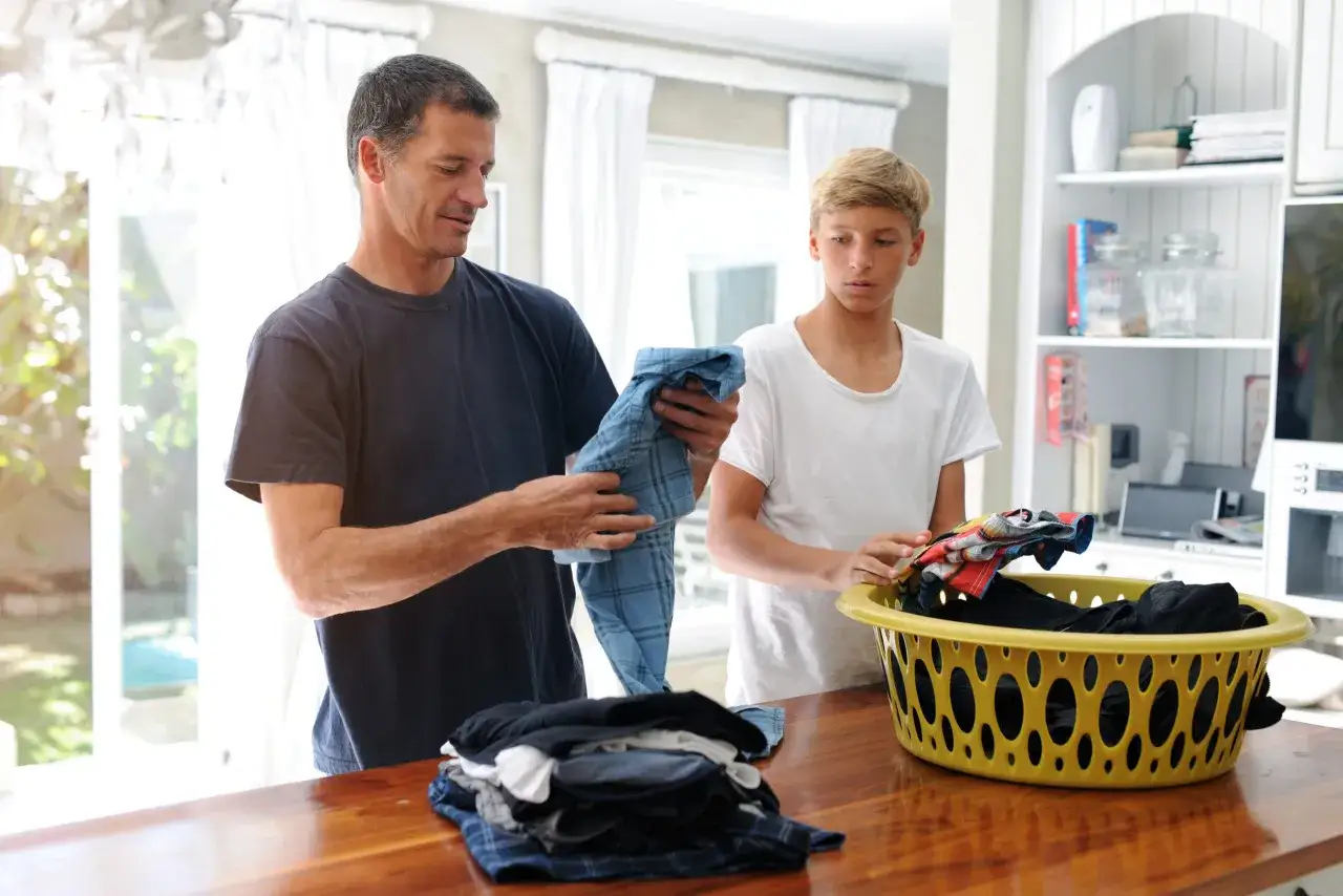Vater und Sohn falten Wäsche. Der Vater hält ein blau kariertes Hemd, der Sohn greift in einen Wäschekorb. Eine kostgeld tabelle ist nicht zu sehen.