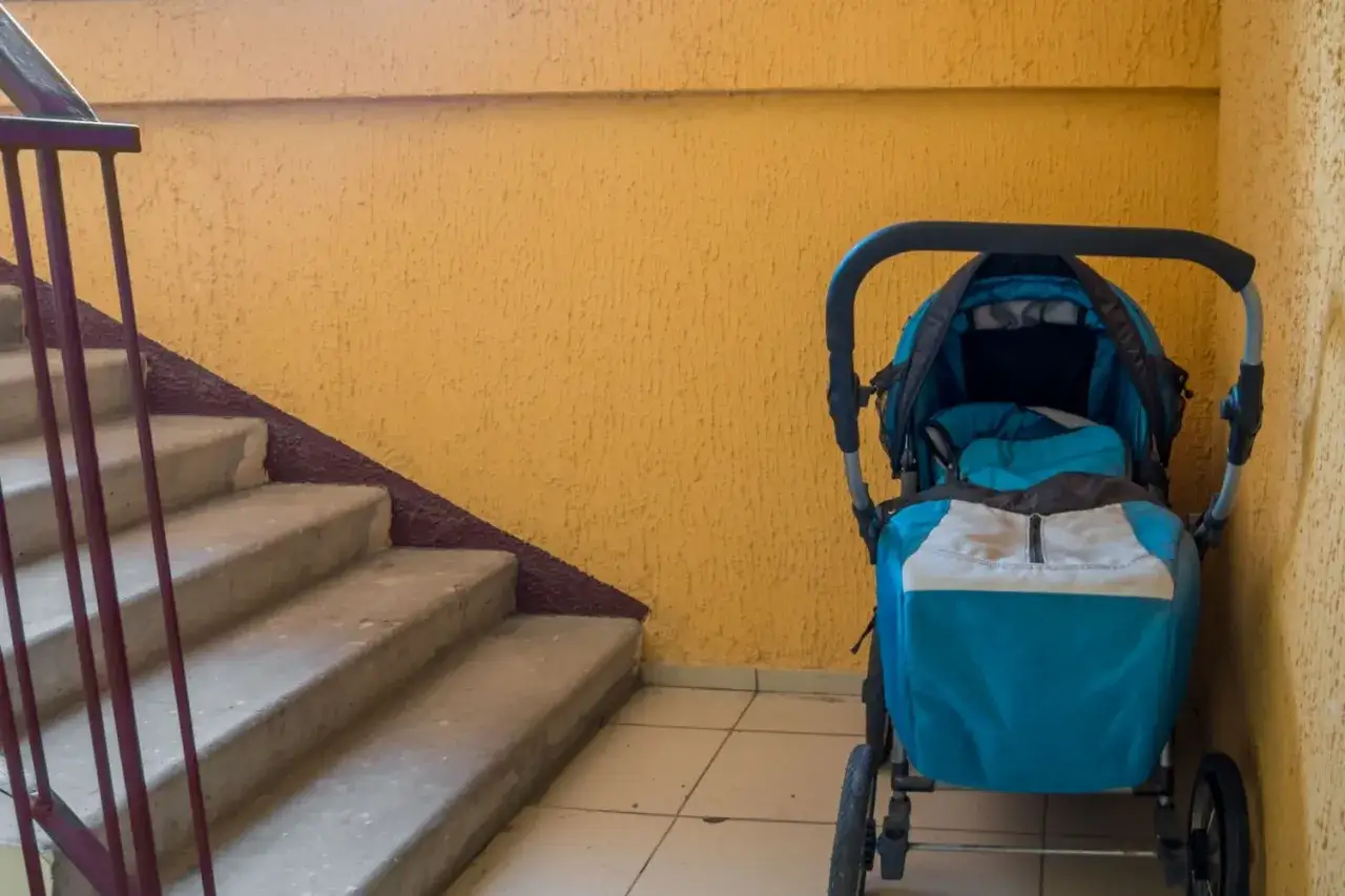 Ein blauer Kinderwagen steht im Treppenhaus neben einer gelben Wand und einer Betontreppe.