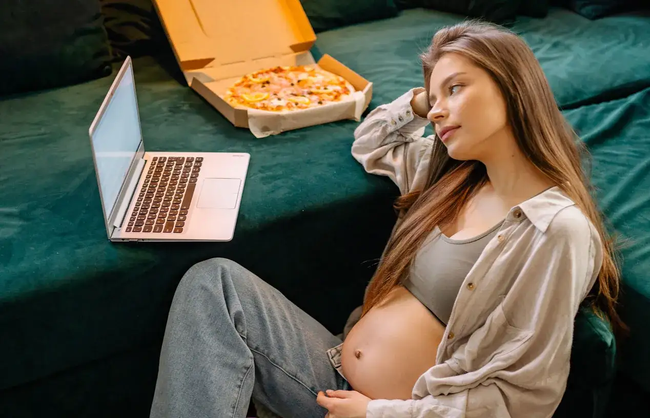 Schwanger im 2. Lehrjahr: Eine junge Frau mit Babybauch lernt am Laptop, während eine Pizza daneben liegt.