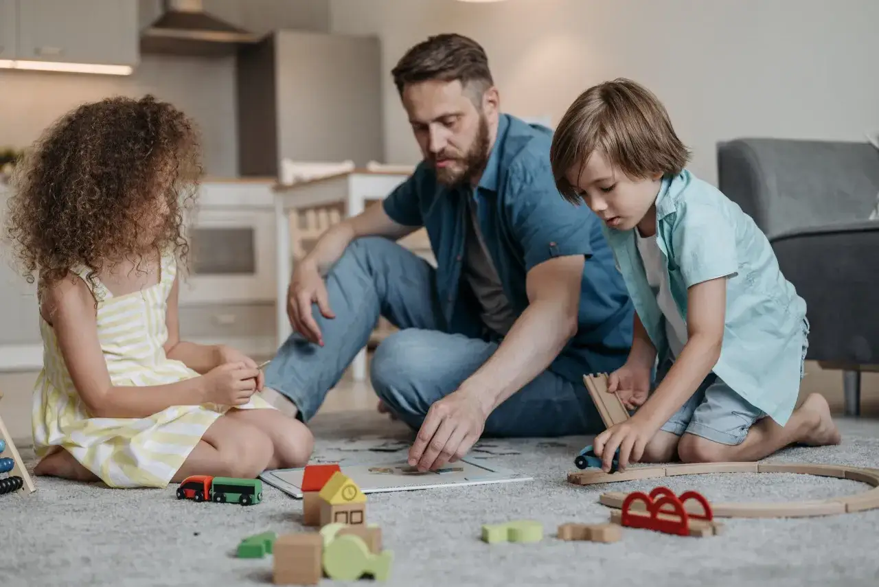 Vater spielt mit Kindern (uvg ab 12 Jahre) auf dem Teppich mit Holzspielzeug.