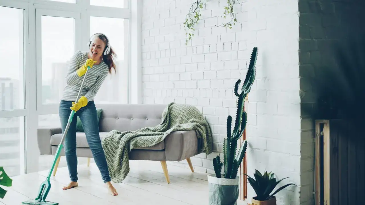 Fröhliche Frau mit Kopfhörern wischt den Boden, als ob sie singt. Das Zimmer ist fast besenrein, nur noch ein paar Pflanzen und ein Sofa.
